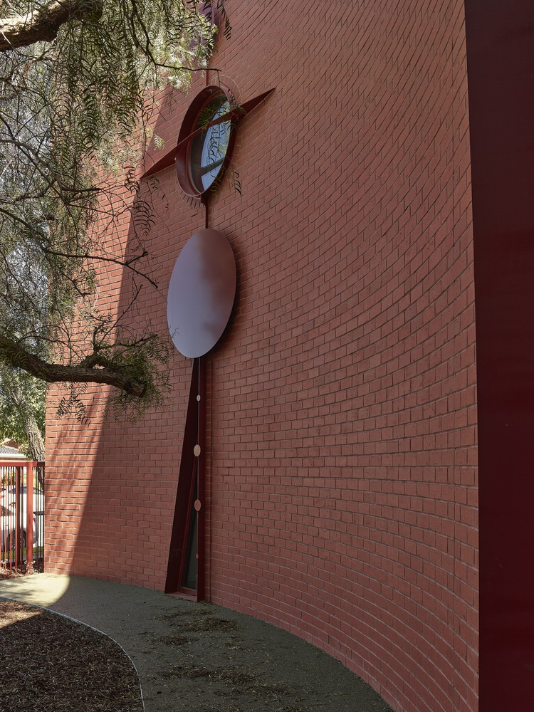 Tall brick wall with vertical metal sculpture featuring circular mirror and oval panels beneath tree branches