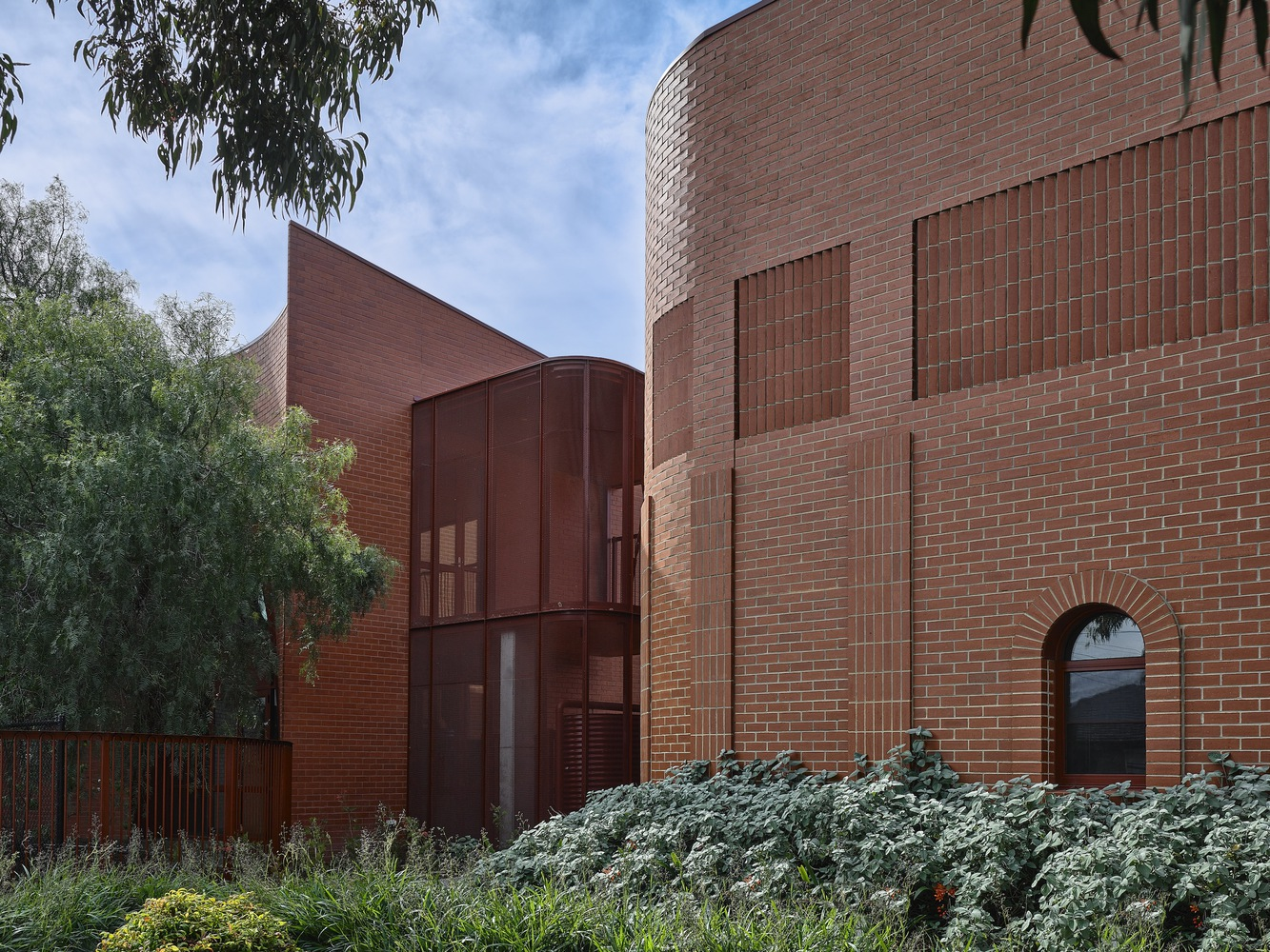 Corner detail of cylindrical brick tower with circular window beside curved metal panel volume and planted foreground