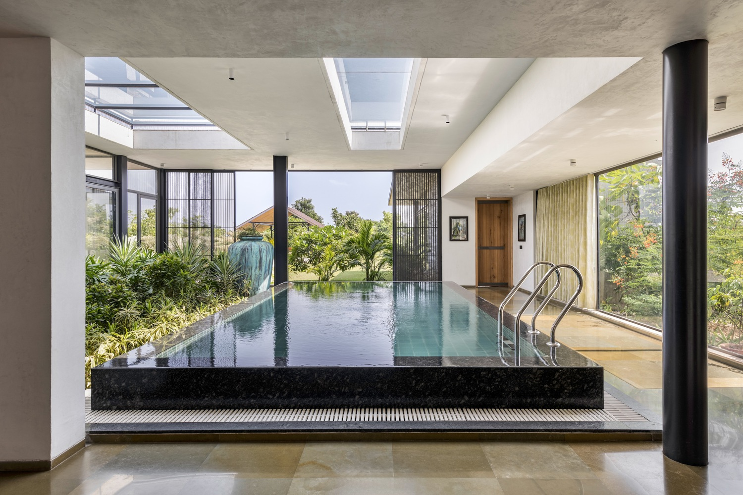 Indoor pool: dark tile pool under a large skylight, black column, full-height glazing to the garden, exposed concrete ceiling