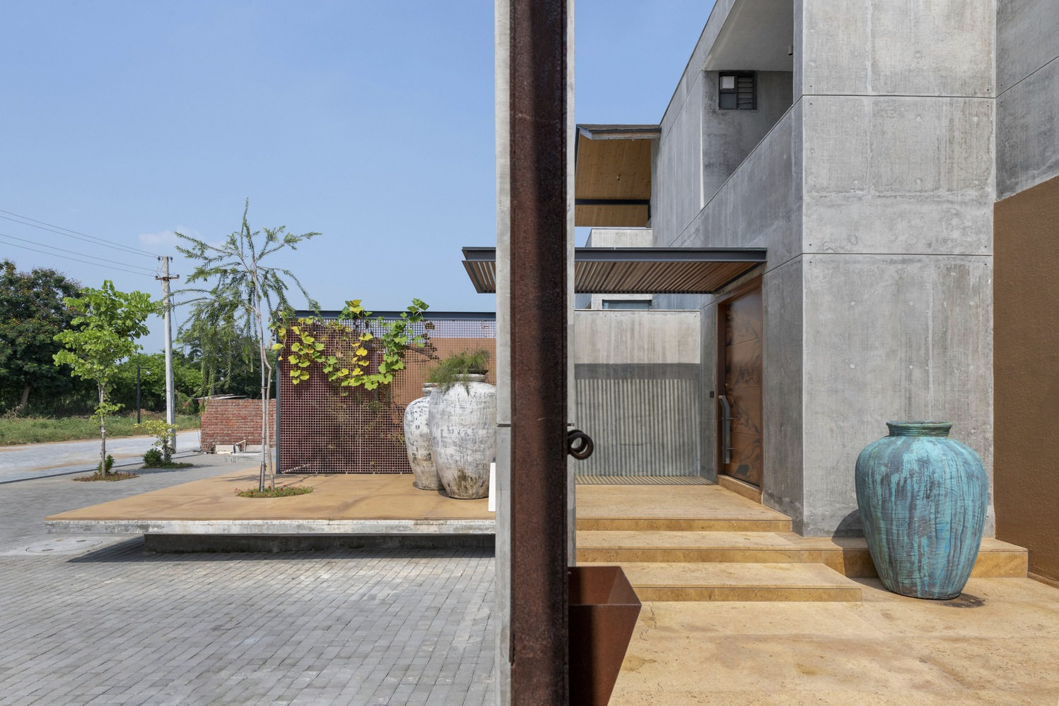 Entry approach: concrete and corten steel volumes, brick paving, young tree, blue ceramic vases, open sky, flat landscape beyond