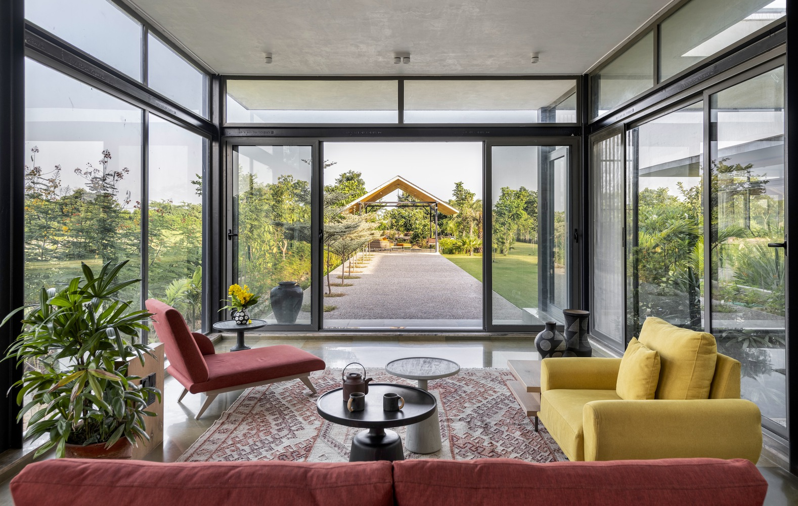 Living room: floor-to-ceiling black steel glazing, red lounge chair, yellow sofa, rug, garden path and timber pavilion visible beyond