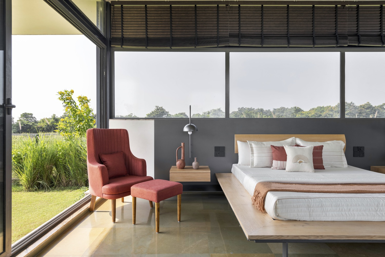 Bedroom: platform bed with white linen, red wingback chair, grey half-wall, full-height glazing open to the lawn, metal louvre ceiling