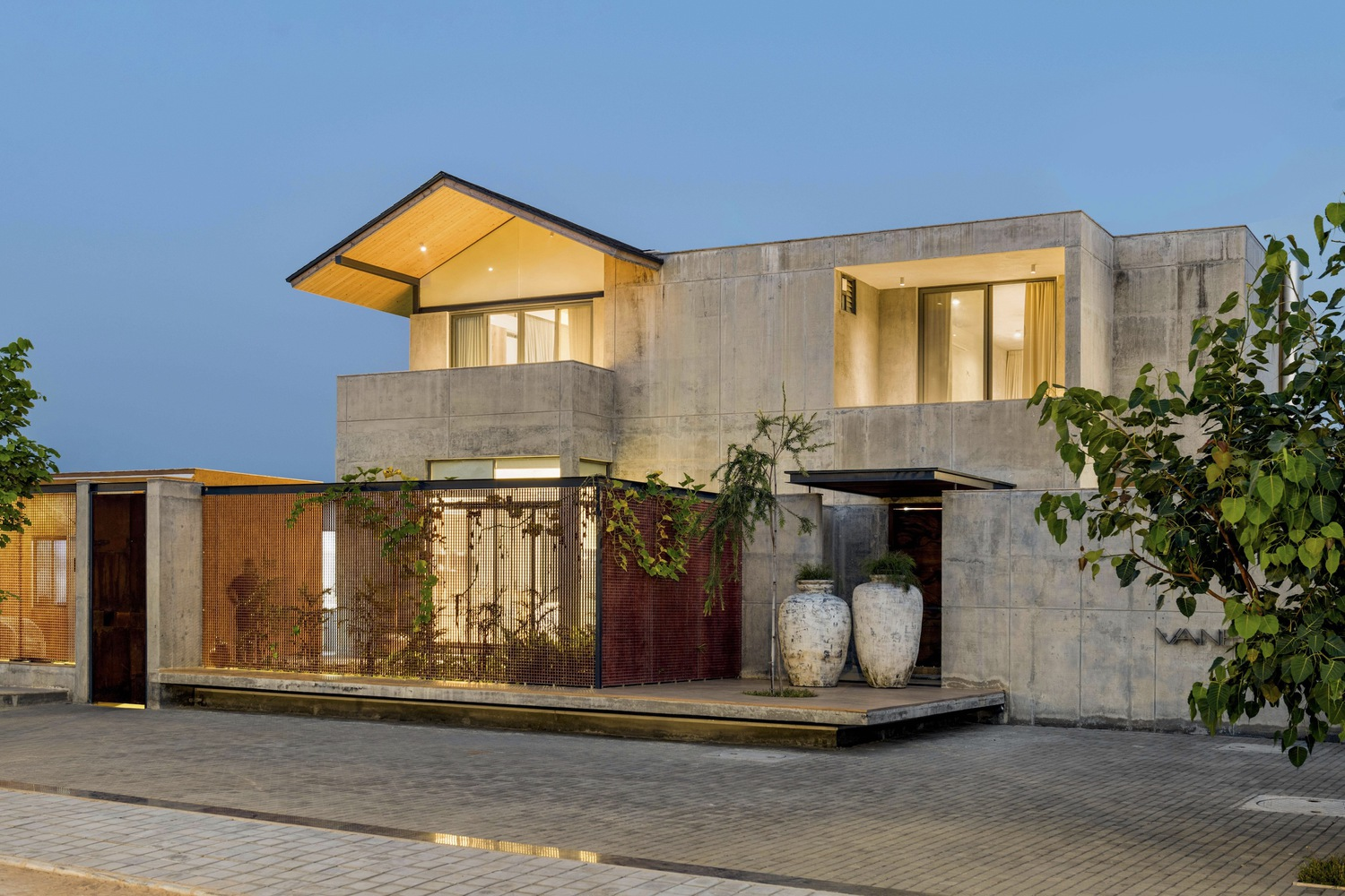 Street facade at dusk: concrete volumes with corten steel gate, pitched roof with warm light glowing from within, timber cladding, blue ceramic vases