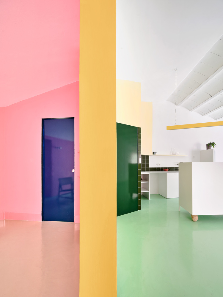 View through yellow column dividing pink floored hallway from green floored kitchen with dark blue door