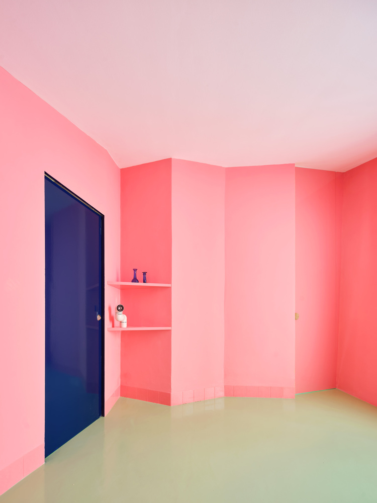 Corner room with pink gradient walls, pale green flooring, corner shelves, and a dark blue door