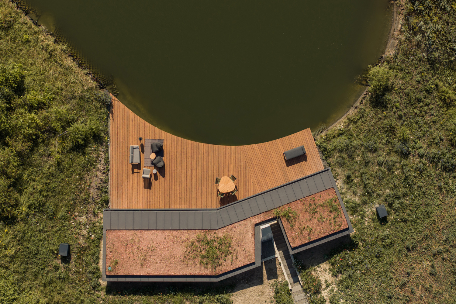 Aerial: V-shaped plan with timber deck wrapping the pond edge, green roof, concrete chimney, meadow surrounding