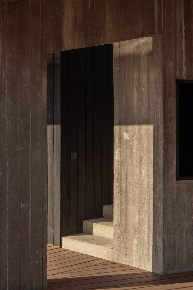 Entry passage: charred timber walls on both sides, concrete step, sharp shadows, timber deck ahead