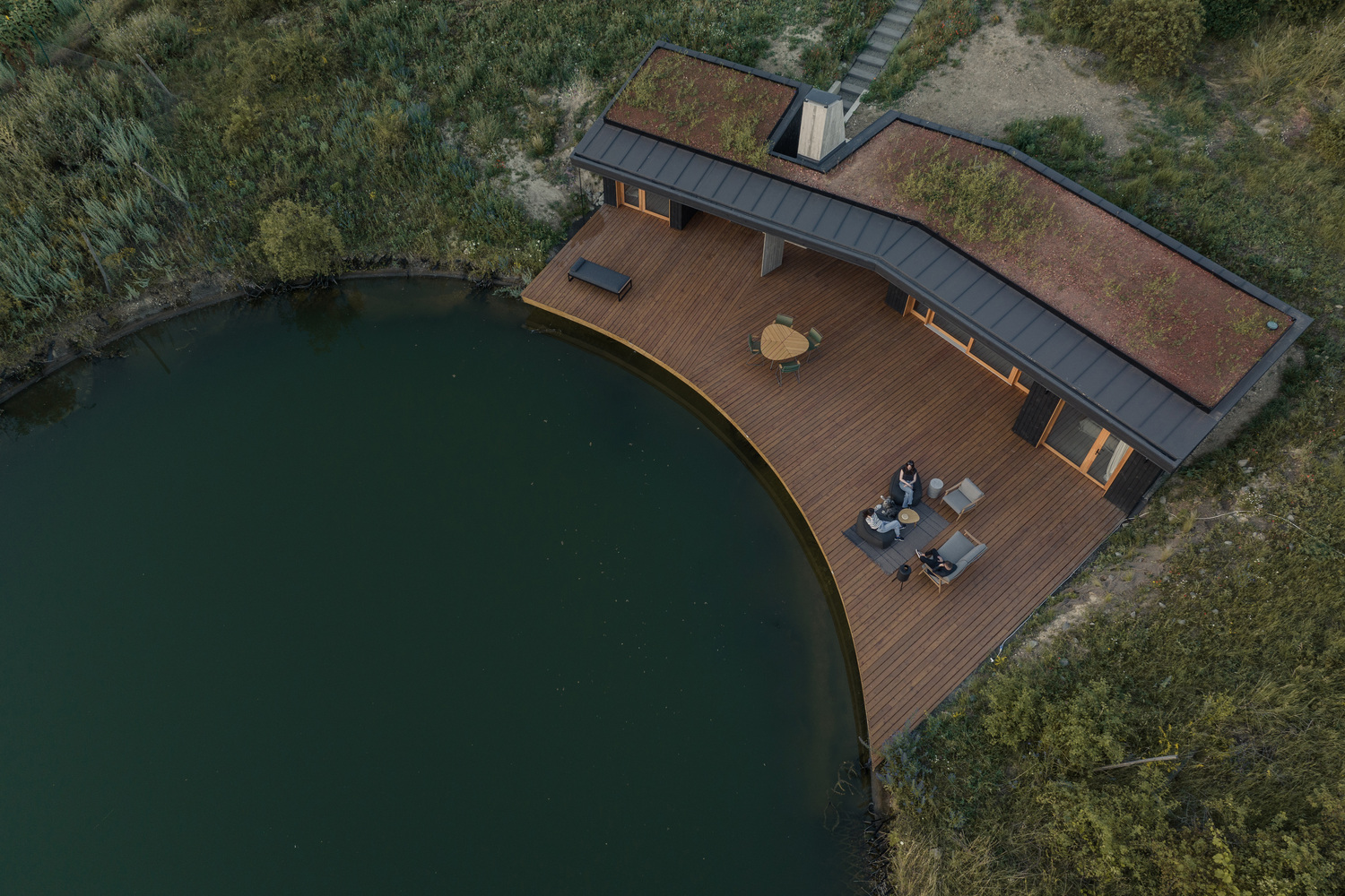 Aerial: V-shaped house with timber deck curving along the pond edge, green roof, two figures sitting on the deck, meadow