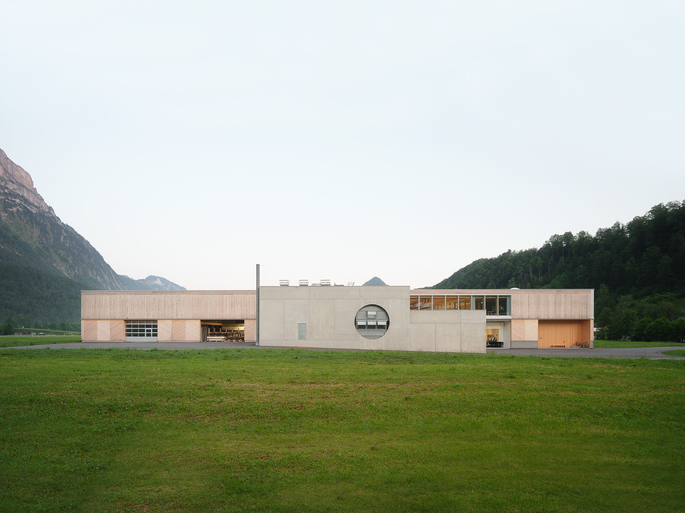 Long horizontal facade combining timber cladding and concrete panels across a grass field with mountains beyond