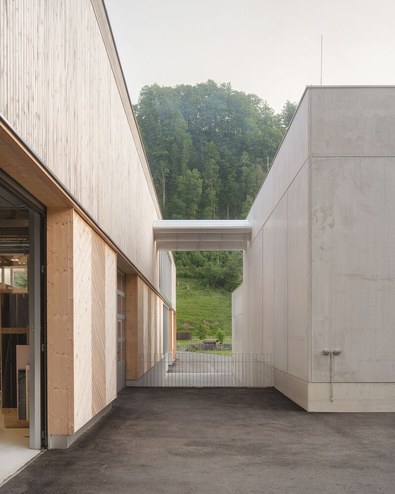 Exterior passageway between timber-clad workshop and concrete volumes framing a view toward forested hills