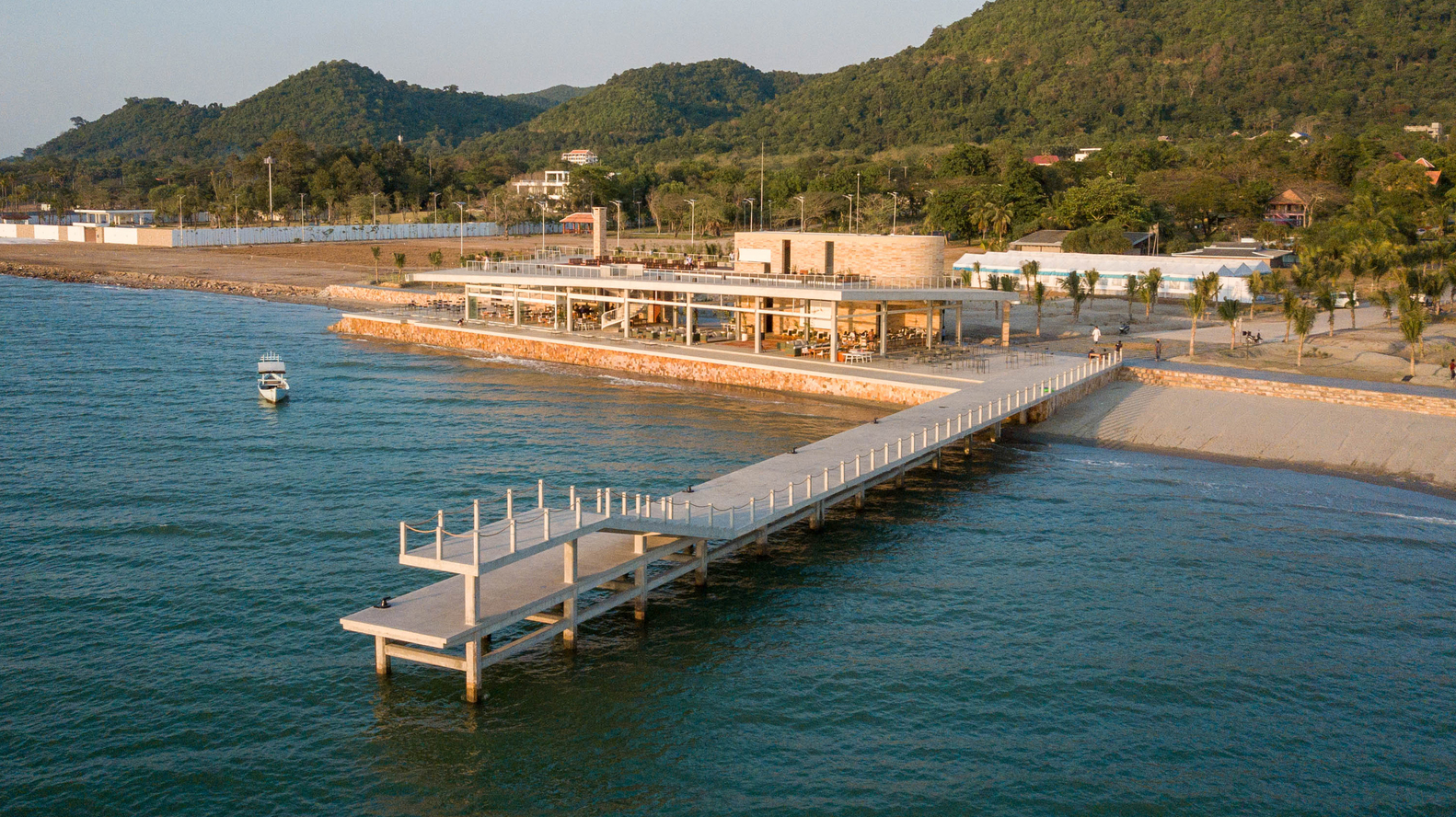 Timber walkway stretching from shore to the glass-walled pavilion at golden hour with forested hills beyond