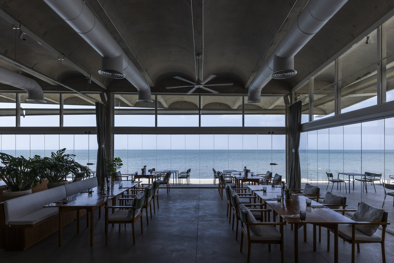 Interior dining space with exposed metal ductwork and floor-to-ceiling windows overlooking the sea