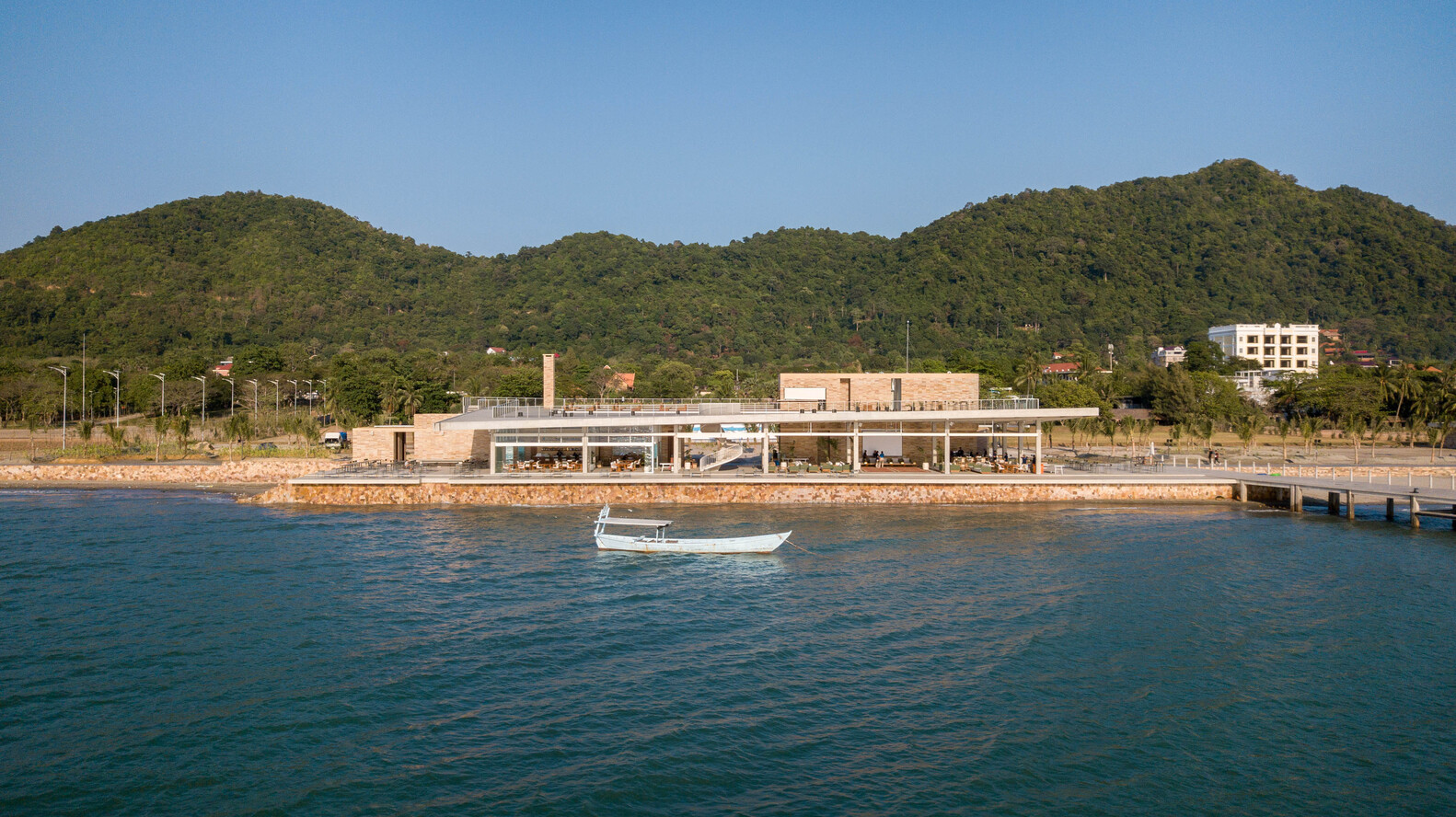 Waterfront view of the low-slung pavilion with exposed concrete columns and brick retaining wall under forested hills