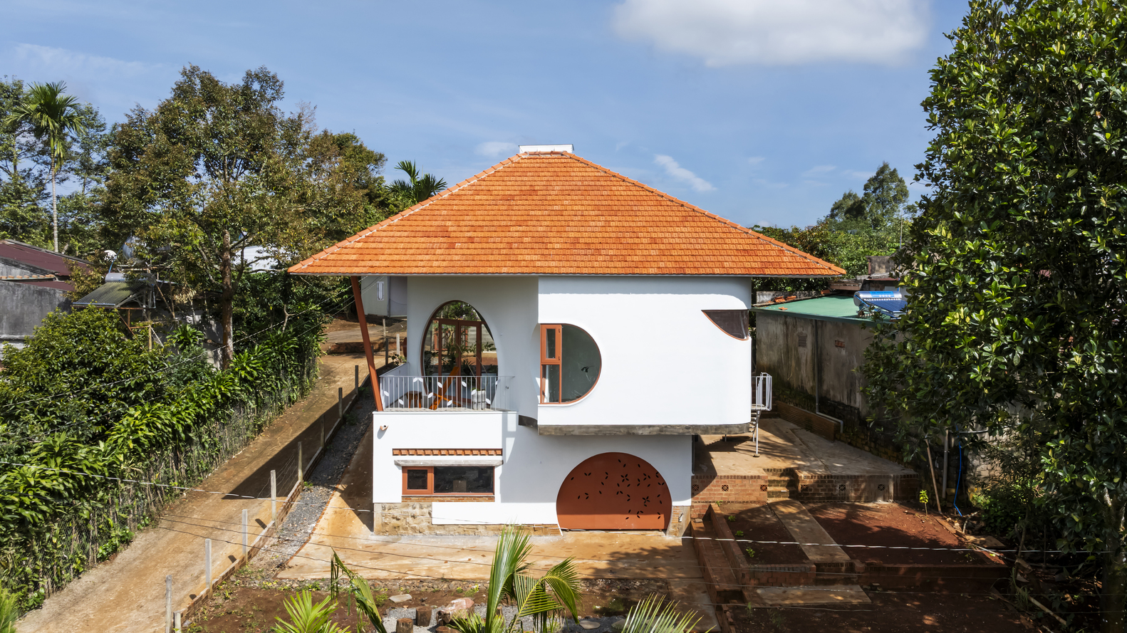 White two-story residence with orange clay tile hipped roof and circular openings set among palm and deciduous trees