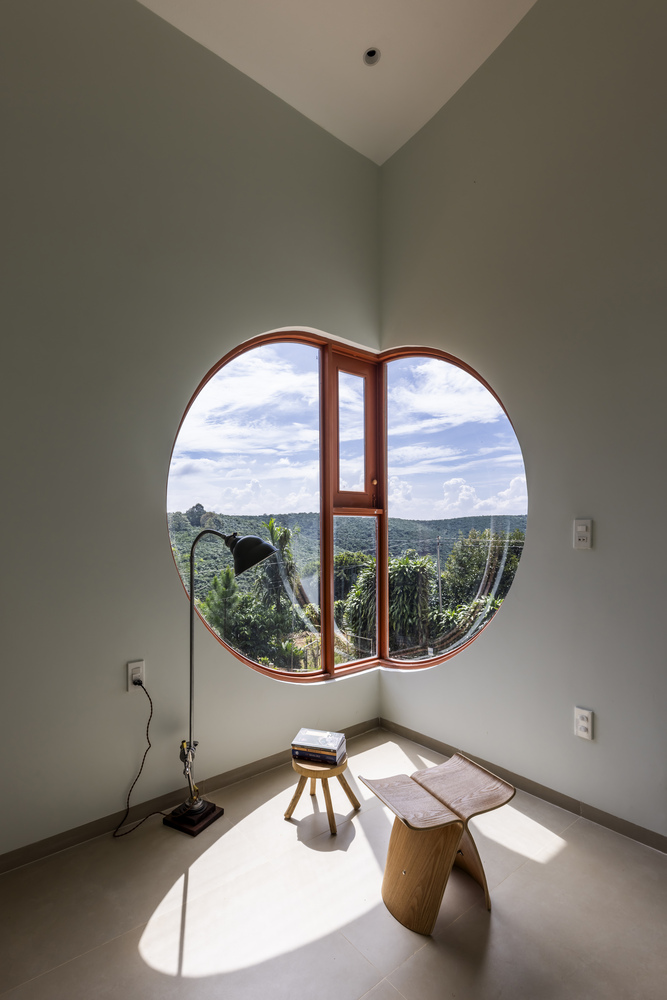 Circular timber window framing rolling green hills under cloudy sky with floor lamp and stool