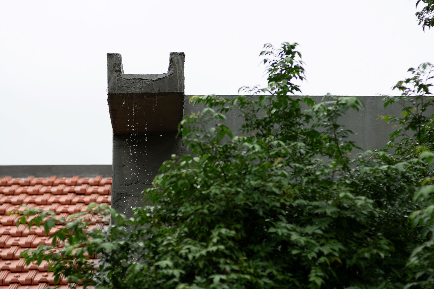 Dark perforated chimney rising above a terracotta tile roof and grey parapet wall with green foliage