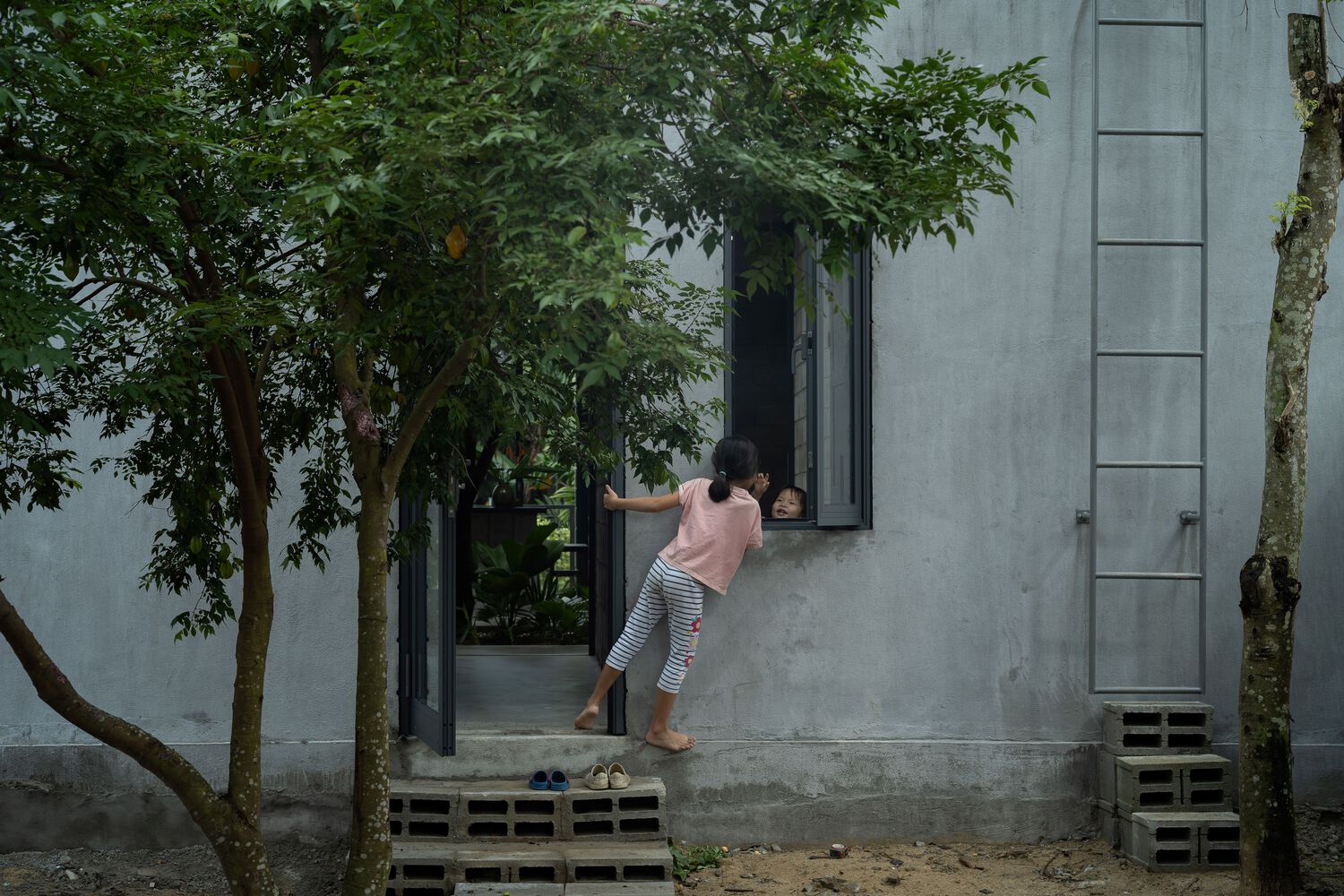 Child climbing through window opening in concrete block facade with exterior ladder and trees