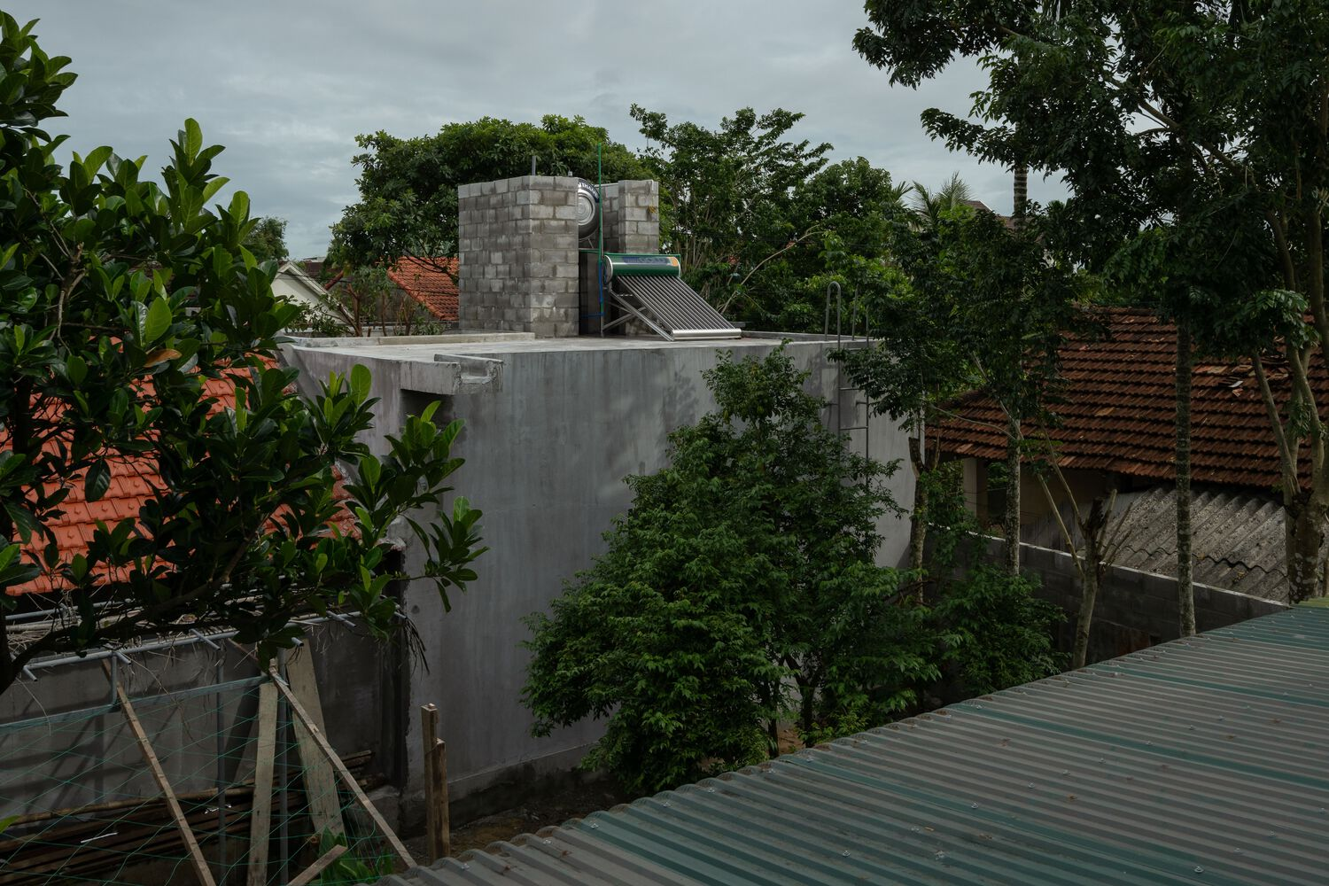 Concrete block volume rising above neighboring tile roofs surrounded by tropical vegetation under overcast skies