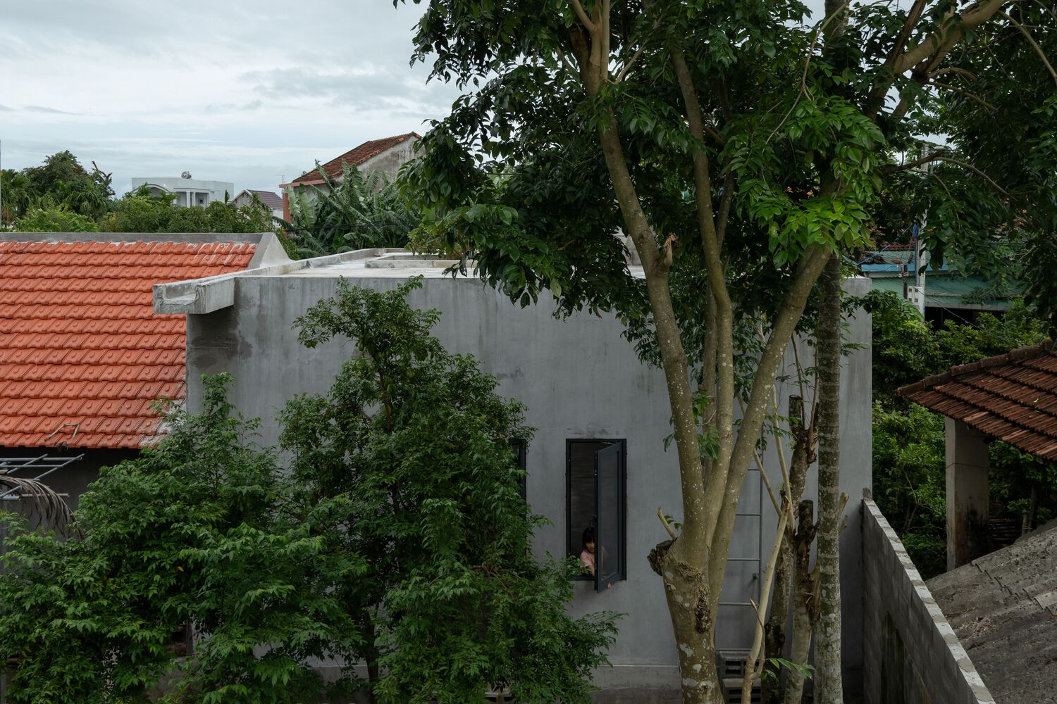Elevated view of the flat concrete roof volume nestled among trees and neighboring tile roofs