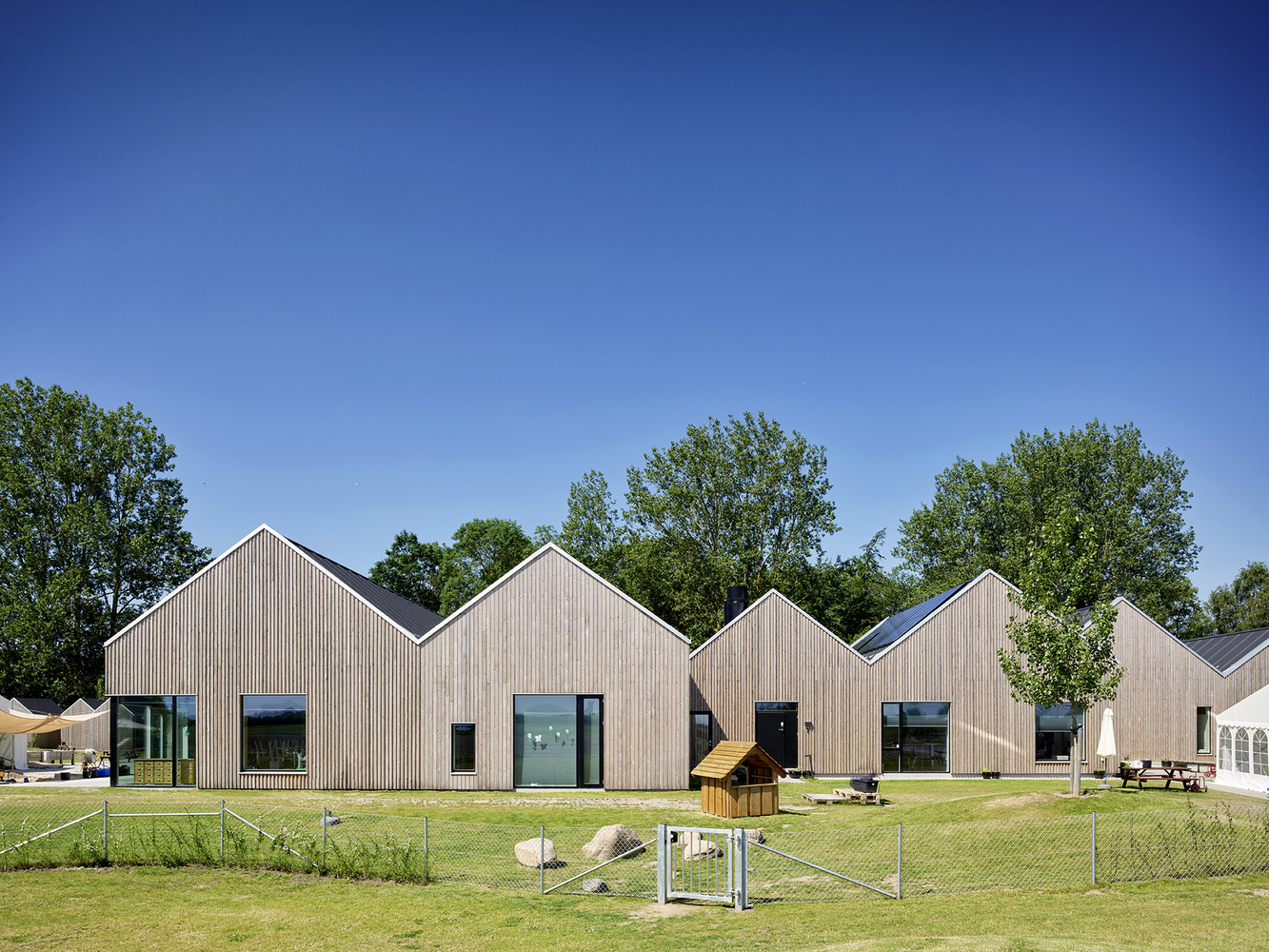 Row of timber-clad gable-roofed volumes fronting a grass lawn with playground equipment and young trees