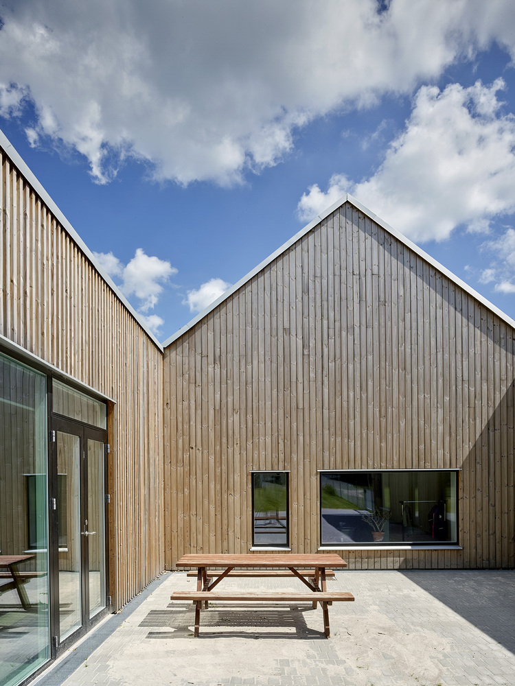 Concrete courtyard with timber picnic benches between vertical timber-clad volumes under scattered clouds