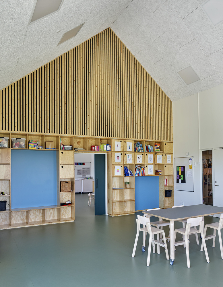 Classroom space with house-shaped storage unit featuring slatted timber gable and built-in plywood cubbies with sliding panels