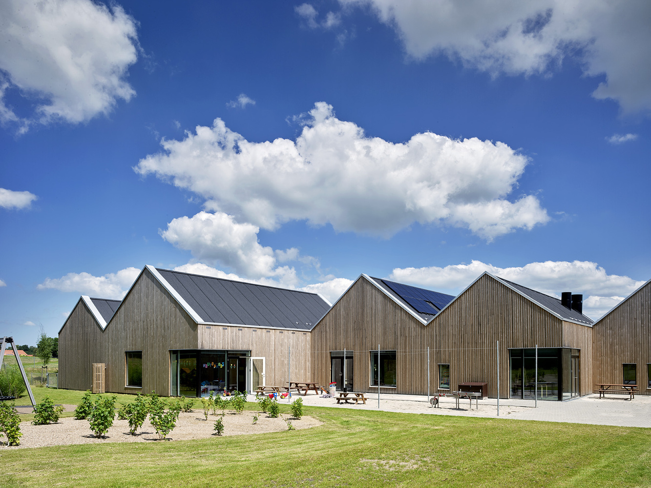Group of linked gabled structures with vertical timber cladding set in open lawn with young plantings