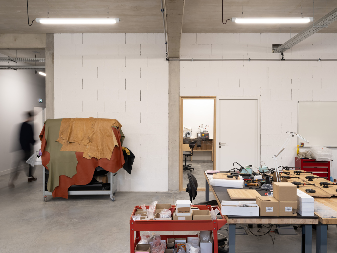 Workshop interior with exposed concrete ceiling, fluorescent tubes, and a person moving past worktables and stored materials