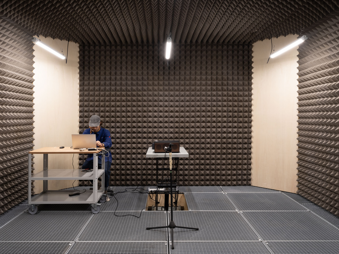 Sound testing chamber lined with pyramidal acoustic foam panels and a person examining equipment at a workbench