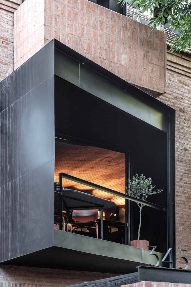 Cantilevered volume clad in dark metal panels with large glazed opening revealing wooden ceiling and dining furniture