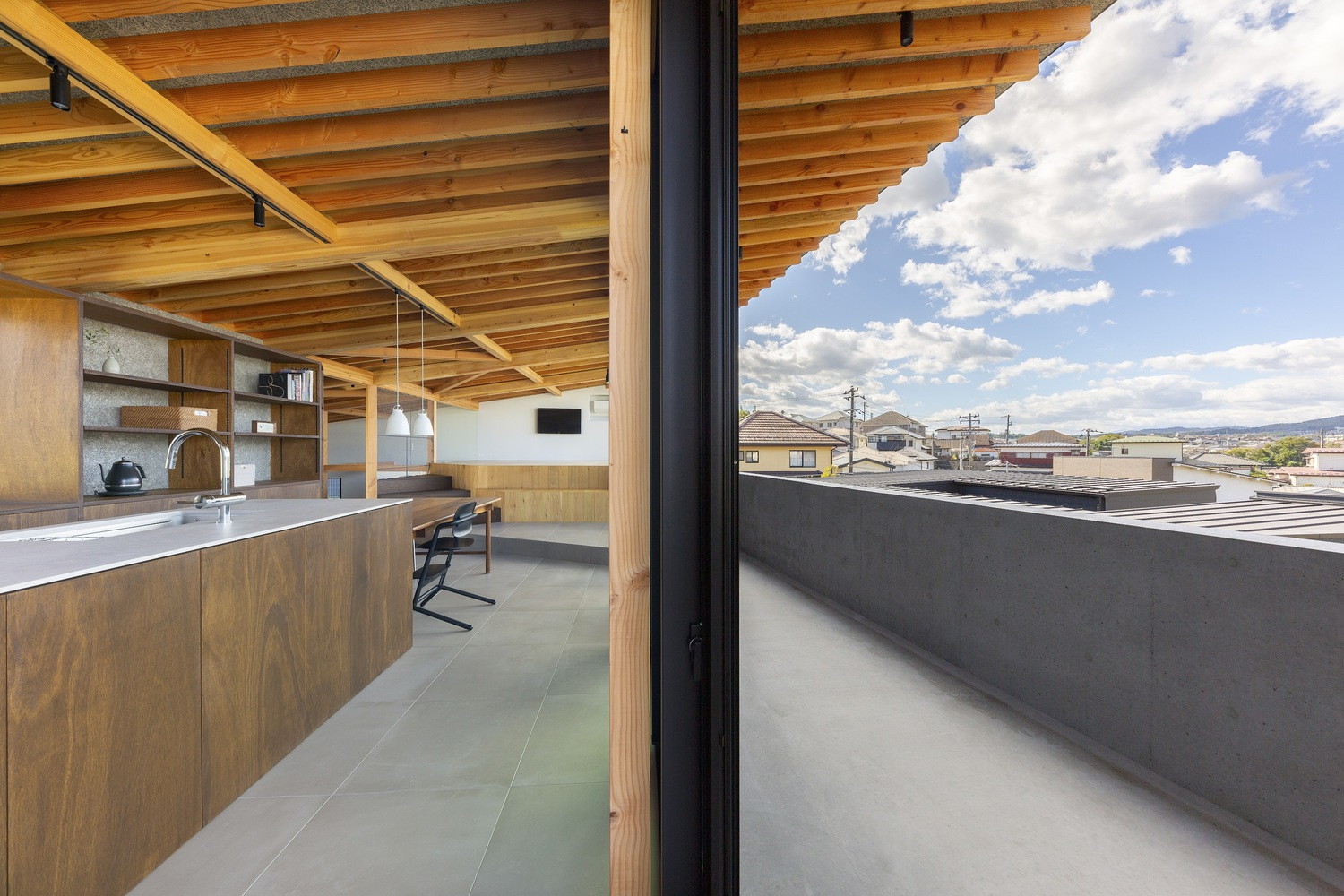 Upper kitchen: exposed timber rafter ceiling, stainless steel counter, dark timber shelving, sliding glass to the balcony with a view across the town rooftops