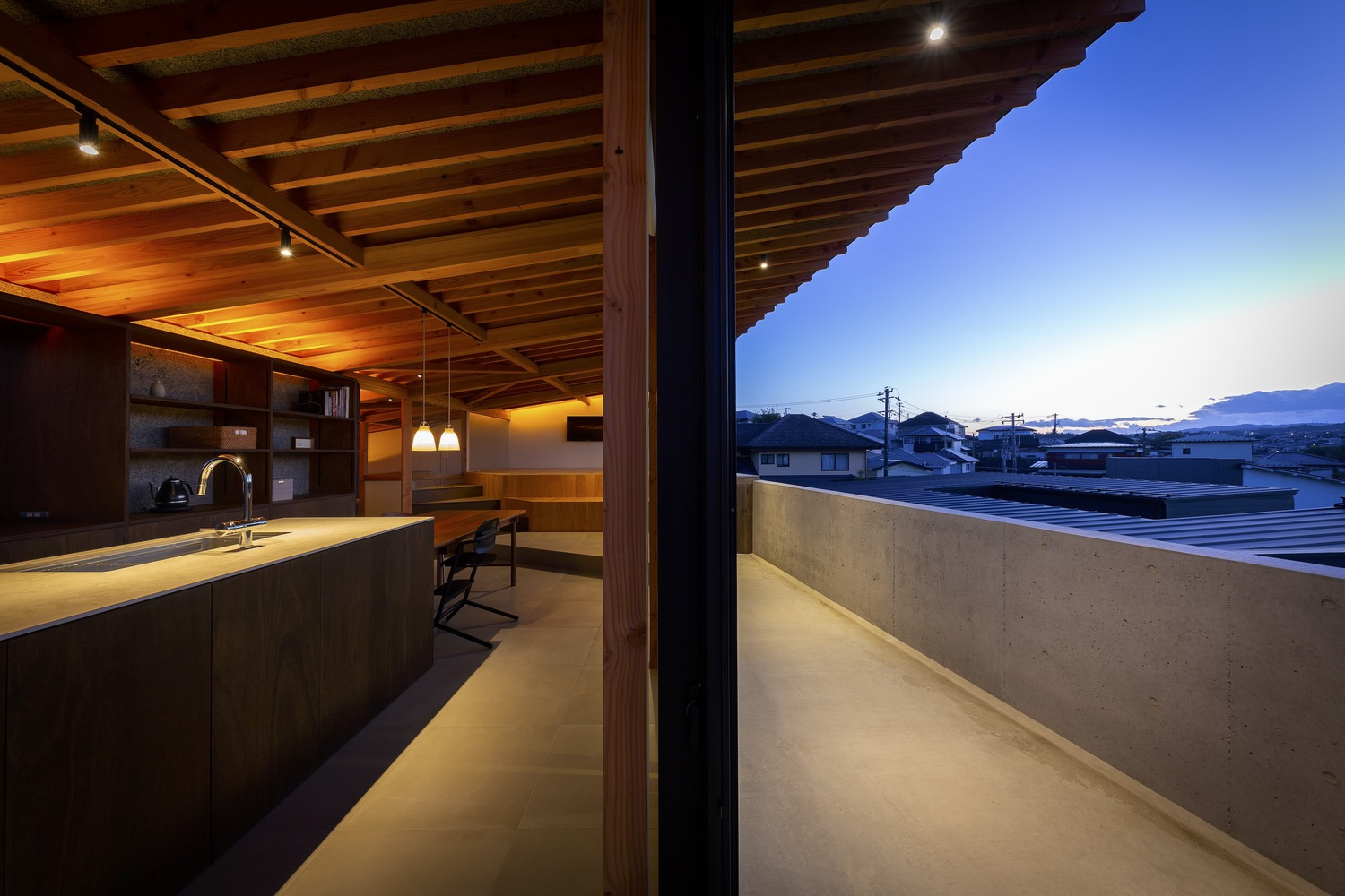 Balcony at dusk: view from the sliding glass onto the concrete deck, warm interior on the left, town and mountains in the blue hour beyond