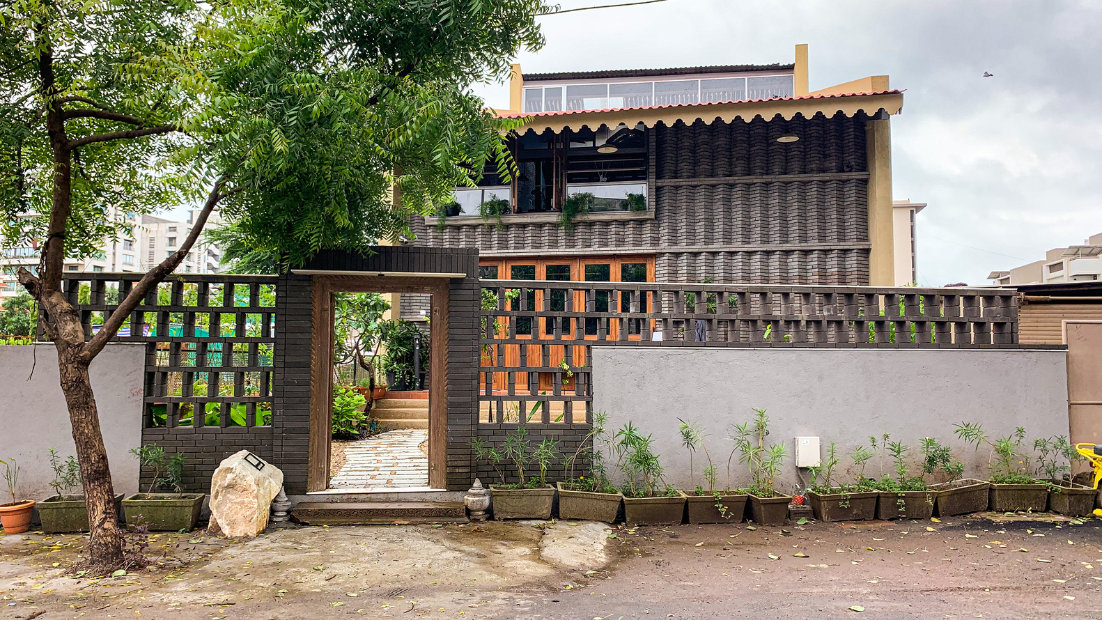 Street facade with perforated brick screen walls, planted bed, and tree beside the entry gate under overcast sky