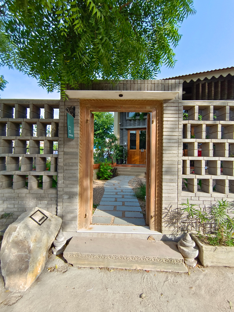 Timber-framed entrance gateway with perforated concrete block walls and a paved pathway under a green tree canopy