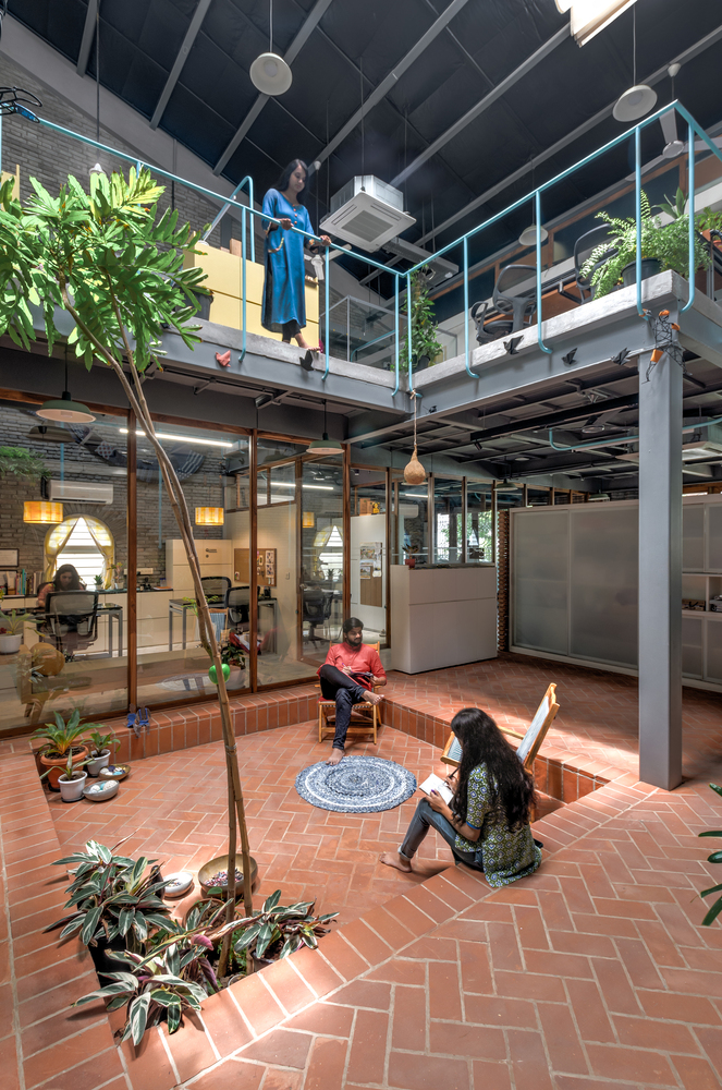 Double-height interior courtyard with terracotta brick floor and people on steel mezzanine walkway with turquoise railings