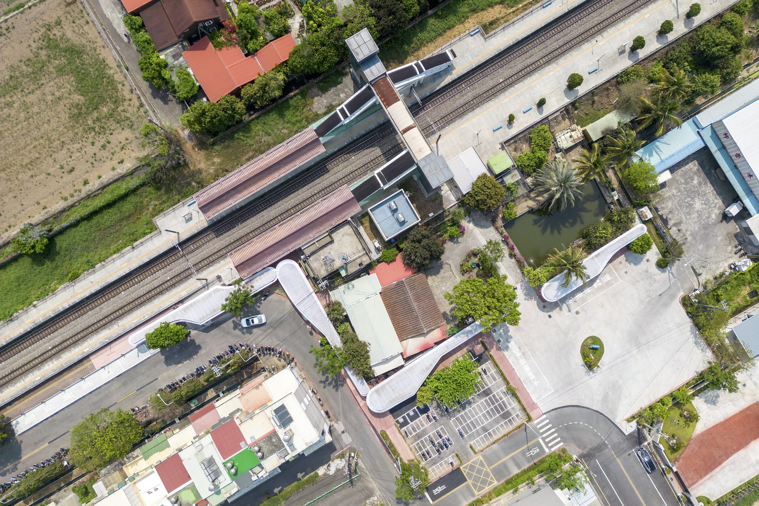 Aerial: curved white canopy beside the railway tracks, red columns, brick low walls, surrounding neighbourhood rooftops