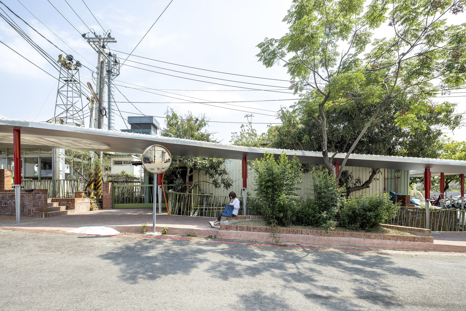Street corner: curved white canopy floating on red columns, brick low wall, trees, figure sitting on steps, overhead wires