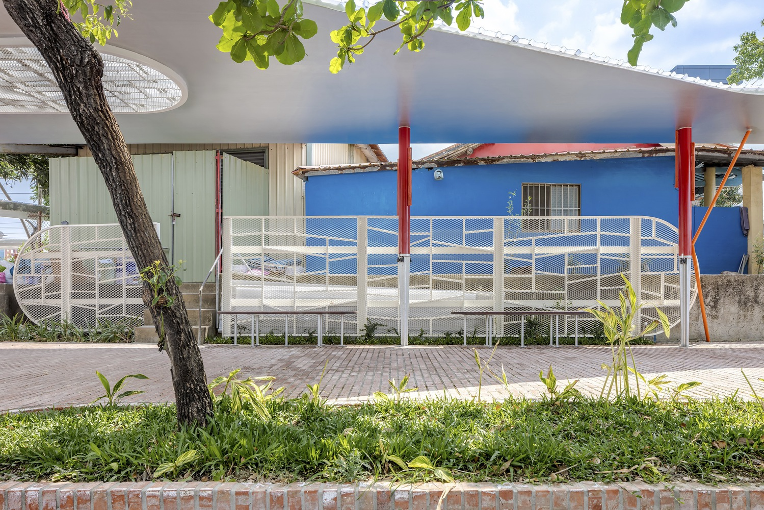 Side view: white canopy over brick paving, red columns, white metal railing, blue building behind, existing trees retained