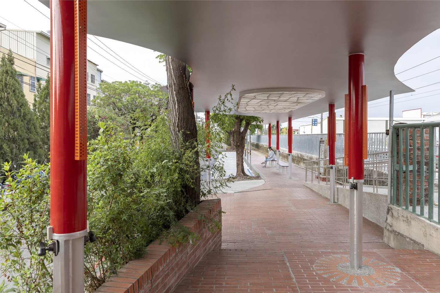 Under the canopy: row of red columns receding in perspective, brick paving, trees and planting at the edge
