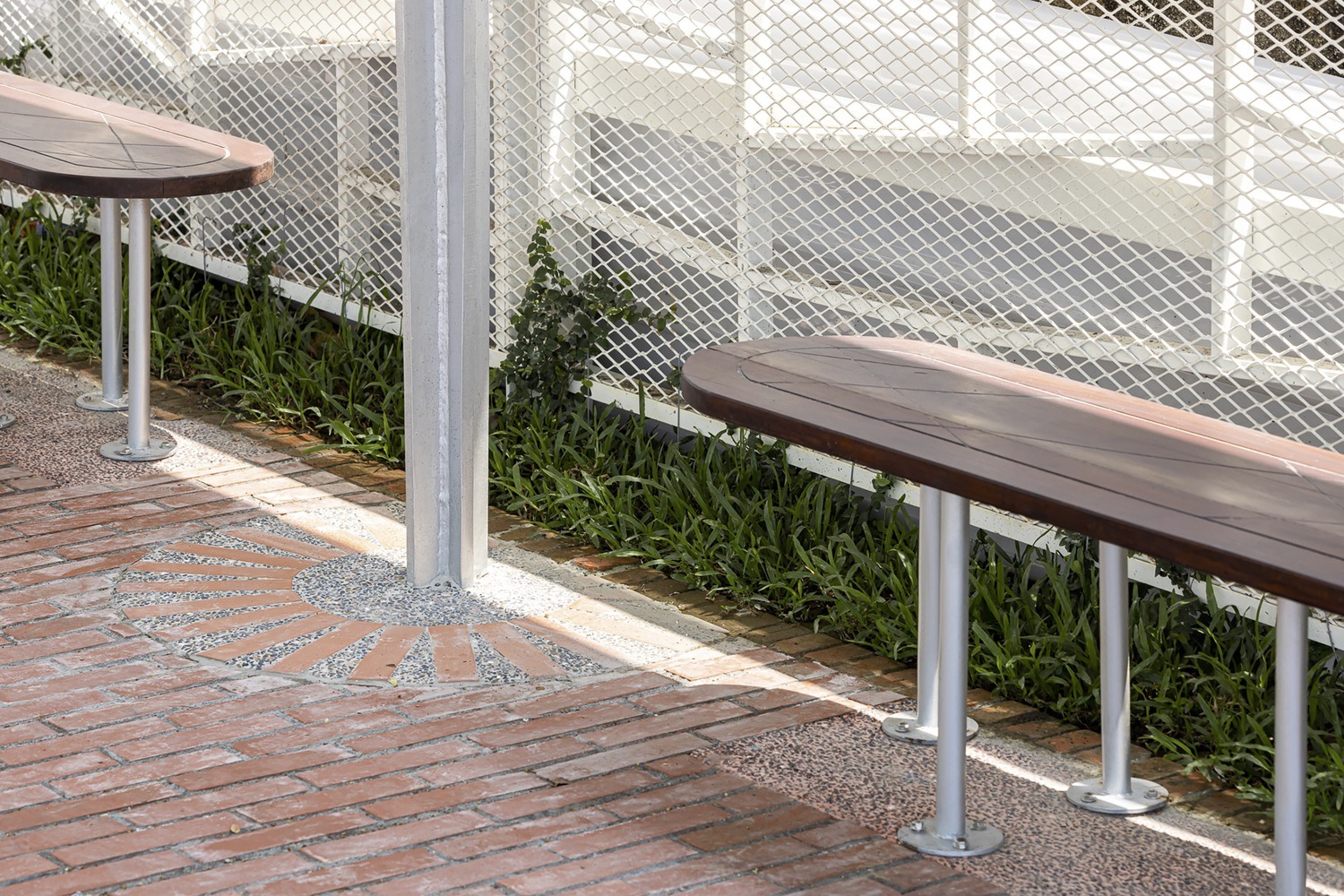 Bench and paving detail: timber-topped benches on steel legs, white mesh railing, green planting strip, radial tile pattern in brick