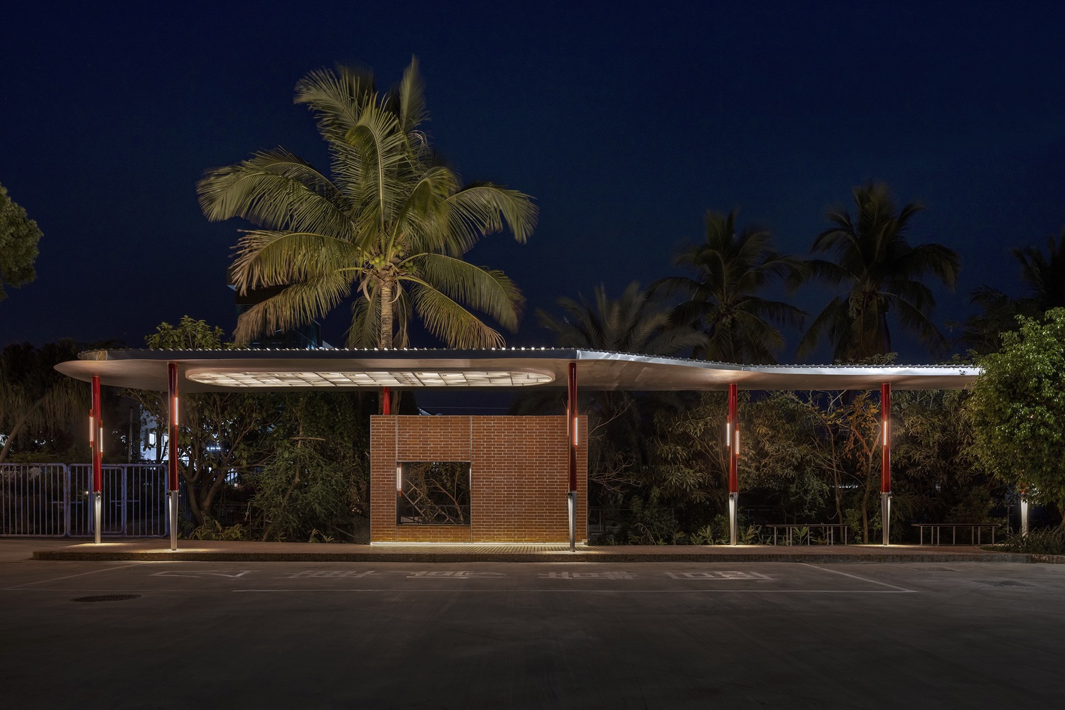 Night from across the street: full pavilion with white canopy floating on red columns, palm tree, brick service volume at centre
