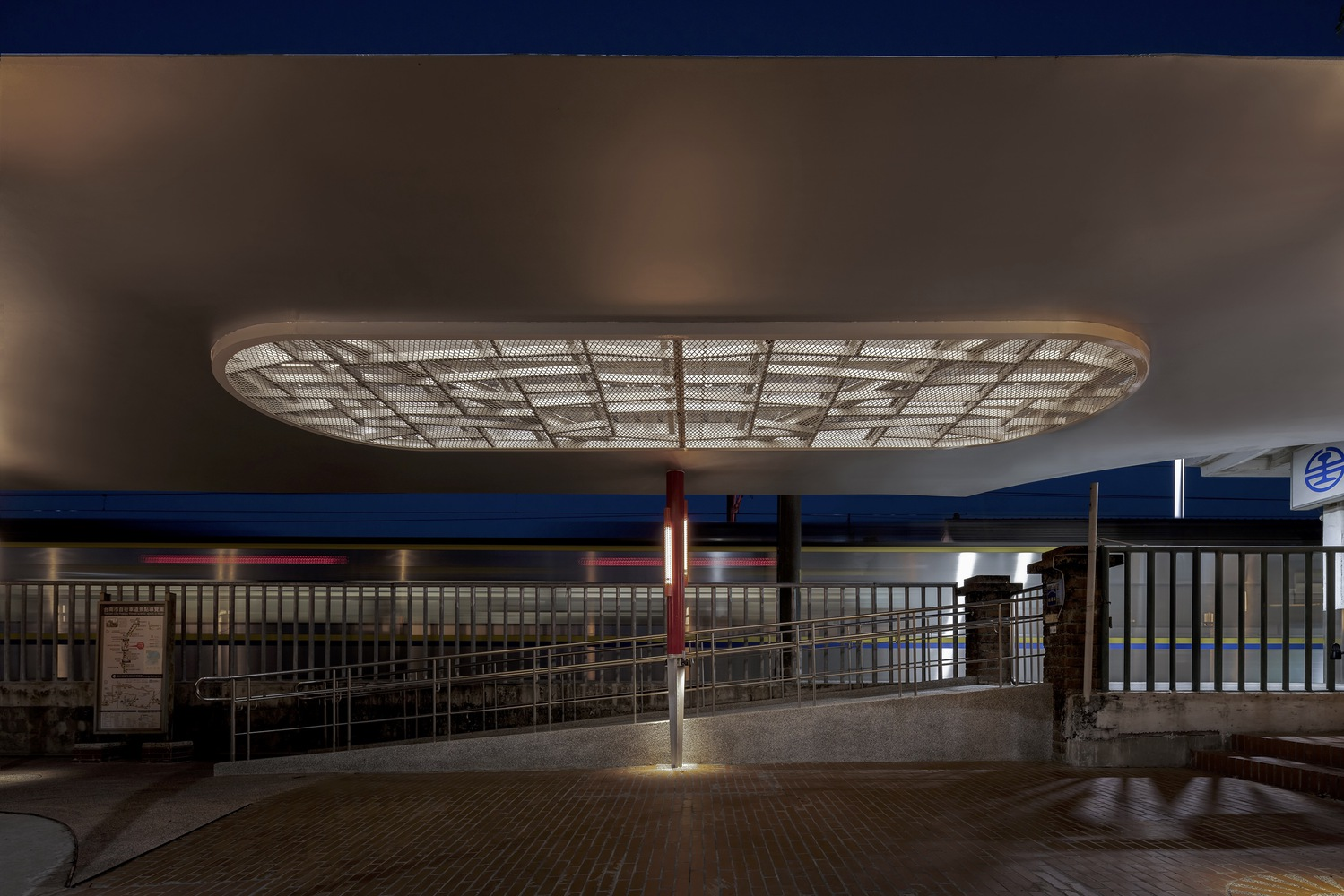 Night: oval skylight lit against blue wall, metal railing, red column, warm glow on the canopy soffit