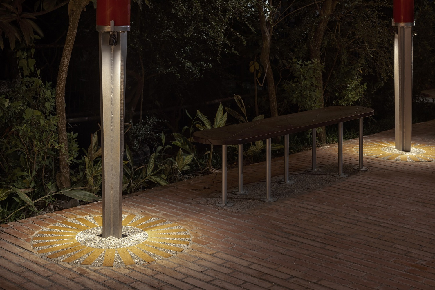 Night detail: red column base with radial sunburst floor light embedded in brick paving, timber bench, planting