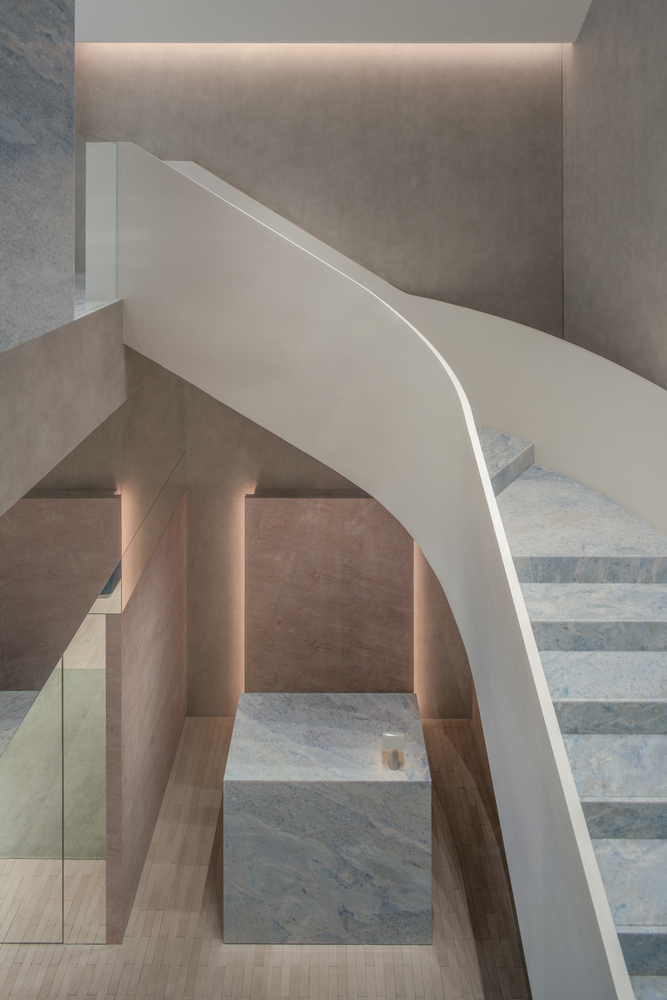 Curved white staircase with marble treads rising between pink stone walls under recessed linear lighting