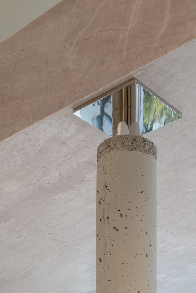 Concrete column topped with a mirrored capital reflecting greenery through a square ceiling opening