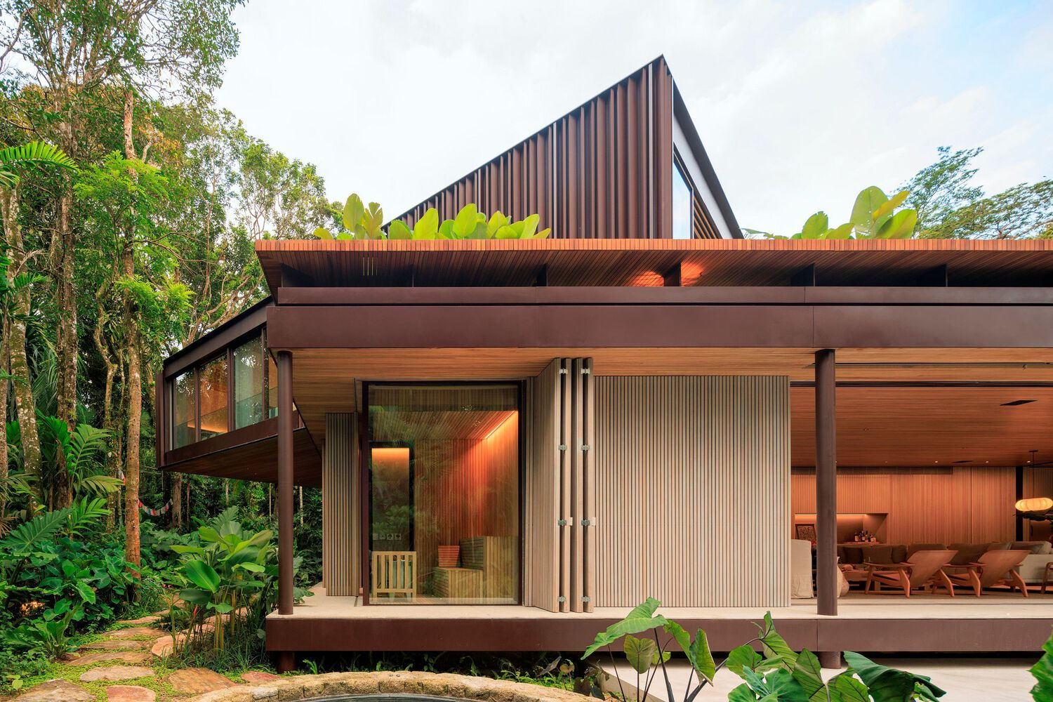 Front facade: two-storey house with dark timber-slatted upper volume, pitched roof, steel columns, covered veranda, tropical garden