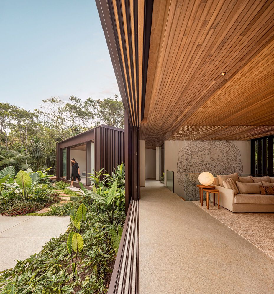 Covered veranda: timber-lined ceiling extending over living area, globe floor lamp, cream sofa, lush Atlantic Forest garden