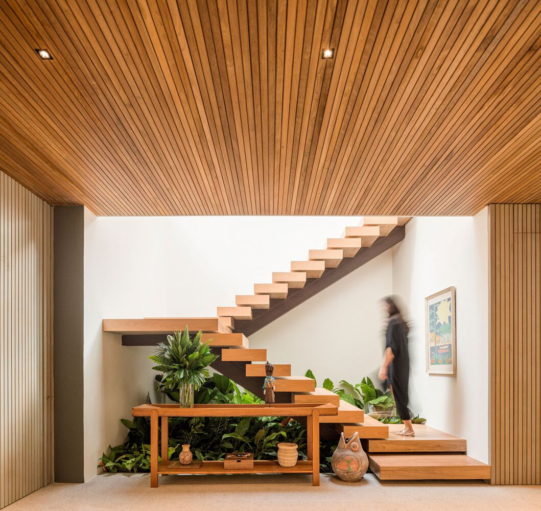 Floating staircase: timber treads cantilevered from white wall, slatted wood ceiling, indoor garden beneath, figure on the stairs