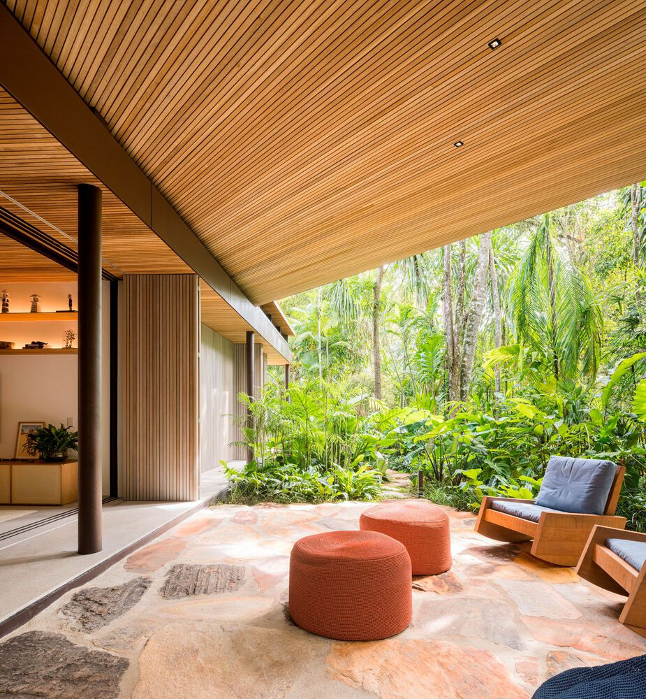Outdoor sitting area: orange poufs and rattan armchair under deep timber ceiling overhang, dense tropical forest beyond