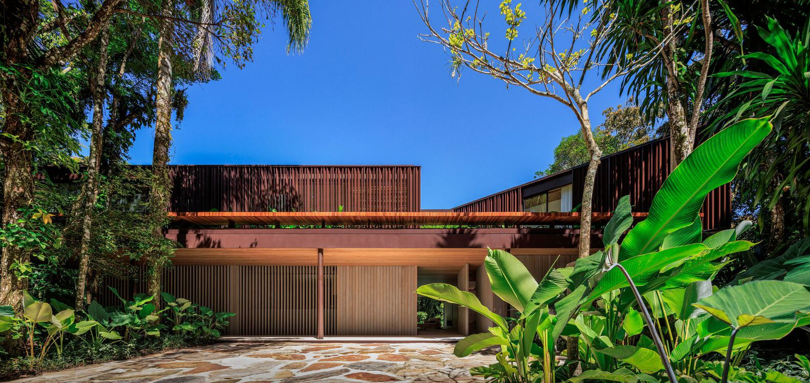 Street facade at dusk: horizontal metal roof slab, timber garage doors, dark-slatted upper volume glowing, tropical planting